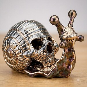 May include: A metallic snail figurine featuring a human skull as its shell. The sculpture has a polished silver finish on the skull portion and a dark, iridescent bronze-toned finish on the snail body, including the two prominent eye stalks.