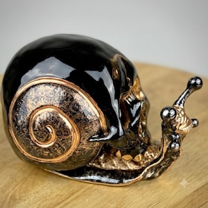 May include: A snail figurine featuring a glossy black shell with a textured bronze spiral pattern. The body of the snail is sculpted to resemble a human skull with metallic bronze accents on the eye stalks and base, creating a dark contrast.