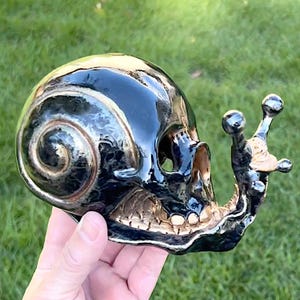 Może przedstawiać: Ceramiczna rzeźba czaszki w kolorze czarnym i złotym z wzorem muszli ślimaka. Czaszka ma szczegółową twarz ze złotymi zębami i muszlę ślimaka na górze głowy. Muszla ślimaka jest czarna z złotymi akcentami.