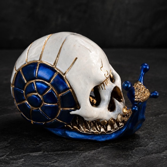A hand-sculpted ceramic snail with a human skull for a shell, finished in striking blue and gold glaze — the kind of numbered, collectible oddity you genuinely can't find anywhere else.