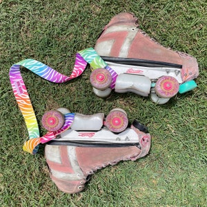 May include: A pair of pink roller skates with white wheels and a rainbow striped strap. The skates have a white base and pink boot with white laces. The strap has the text "EBB & FLO" printed on it.