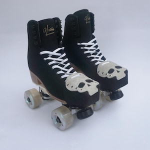 May include: A pair of black roller skates with white laces and light gray suede toe guards featuring a skull design. The boots are labeled Vista by Jackson in gold lettering on the tongue. The skates have clear wheels and metal trucks.