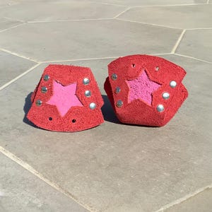 Puede incluir: Dos muñequeras de cuero rojo con un recorte de estrella rosa y remaches plateados. Las muñequeras están hechas de un material texturizado y están diseñadas para envolverse alrededor de la muñeca.