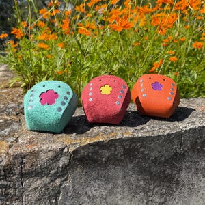 May include: Three colorful, cube-shaped objects with flower cutouts and silver studs are displayed on a stone surface. The objects are teal, red, and orange, with pink, yellow, and purple flower accents. Orange flowers bloom in the background.