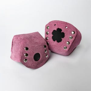 Puede incluir: Dos muñequeras de ante rosa con tachuelas plateadas. Una tiene un recorte en forma de flor que revela un interior negro. La otra tiene una abertura circular. Ambas están diseñadas para envolver la muñeca.