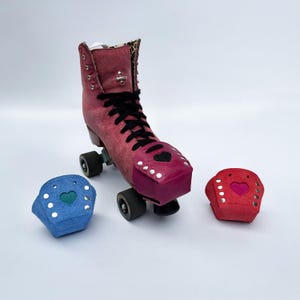 Könnte beinhalten: Ein Rollschuh aus rosa Wildleder mit schwarzen Schnürsenkeln, silbernen Nieten und einem Innenfutter mit Leopardenmuster. Der Rollschuh hat eine rosa Zehenkappe mit einem schwarzen Herz-Ausschnitt. Zwei weitere Zehenkappen sind enthalten: eine blaue mit einem grünen Herz und eine rote mit einem rosa Herz.