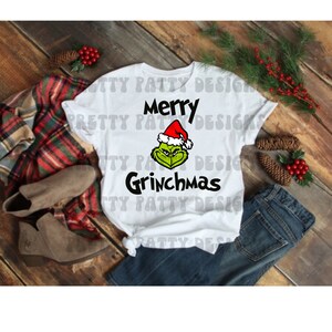 Può includere: Maglietta bianca con la scritta "Merry Grinchmas" e un Grinch verde con un cappello da Babbo Natale.