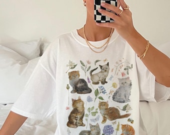 May include: White t-shirt featuring a watercolor illustration of various cat breeds among flowers and foliage. The design includes cats in shades of brown, gray, and orange, with blue and purple floral accents. The shirt has a relaxed fit.
