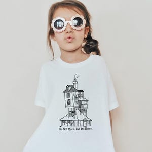 May include: A white t-shirt featuring a detailed black and white illustration of a multi-story house with a chimney. The text "It's Not Much, But It's Home" is printed below the house. The shirt is worn by a child wearing sunglasses.