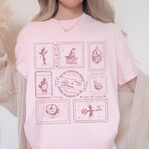 May include: A light pink t-shirt featuring a collection of postage stamp designs in a darker pink hue. The stamps depict various travel-themed illustrations, including a fairy, a wizard's hat, and a key. The words "Air Mail Travel" are also visible.