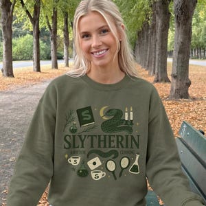 May include: An olive green crewneck sweatshirt featuring a graphic print with the text SLYTHERIN, AMBITION, and CUNNING. The design includes illustrations of a snake, a book, candles, potions, a mirror, a letter, and cups in shades of green and cream.