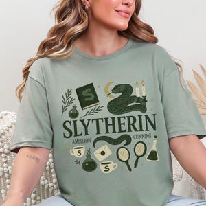 May include: Sage green t-shirt with a Slytherin design. The graphic includes a snake, candles, a book, and the words "Slytherin, Ambition, Cunning." The design is in shades of green, gold, and white.