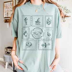 May include: A light green t-shirt featuring a collection of postage stamp designs. The stamps depict various images, including a wizard's hat, a castle, and a teacup. The word "TRAVEL" is printed on one of the stamps.