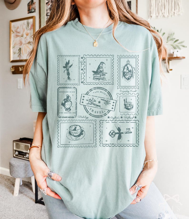 May include: A light green t-shirt featuring a collection of postage stamp designs. The stamps depict various images, including a wizard's hat, a castle, and a teacup. The word "TRAVEL" is printed on one of the stamps.