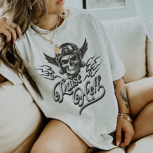 以下が含まれることがあります： 翼の生えたバイクヘルメットをかぶった骸骨と「Raise Hell」の文字が黒で描かれた、ライトグレーのTシャツ。 ゆったりとしたオーバーサイズのフィット感。 デザインは黒です。