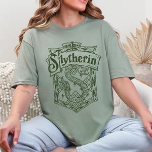 May include: Sage green t-shirt featuring a dark green Slytherin crest. The crest includes a snake, the word "Slytherin" in a banner, and geometric patterns. The shirt is a relaxed fit.