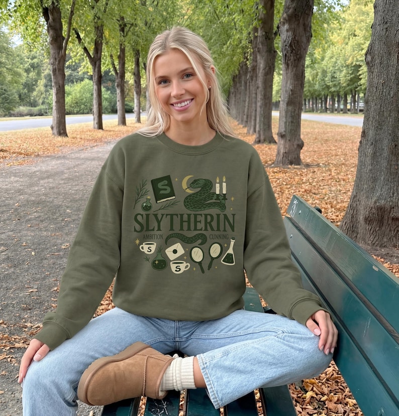 May include: An olive green crewneck sweatshirt featuring a graphic print with the text SLYTHERIN, AMBITION, and CUNNING. The design includes illustrations of a snake, a book, candles, potions, a mirror, a letter, and cups in shades of green and cream.