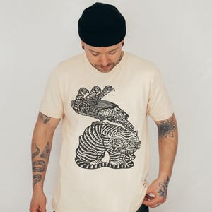 May include: Cream-colored t-shirt featuring a black illustration of a bird with three heads perched atop a tiger. The tiger has black stripes and is in a seated position. The person is wearing a black beanie.