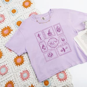 May include: A lilac t-shirt with a series of postage stamp designs in a darker shade of purple. The stamps showcase whimsical illustrations, such as a fairy, a witch's hat, and a key. The text "Air Mail Travel" is also visible.