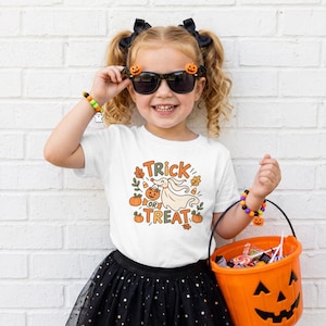 Peut inclure: Un t-shirt blanc avec un motif de chien fantôme tenant un seau citrouille et le texte Trick or Treat, entouré de citrouilles, de feuilles et de bonbons. Le t-shirt est porté avec une jupe noire, des lunettes de soleil et des bracelets de perles.