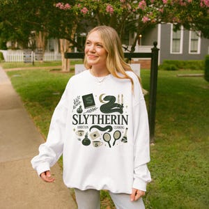 May include: A white long-sleeve sweatshirt featuring a dark green graphic print with the text SLYTHERIN, AMBITION, and CUNNING. The design includes illustrations of a snake, a book with an S, candles, potions, a mirror, and a coffee cup.