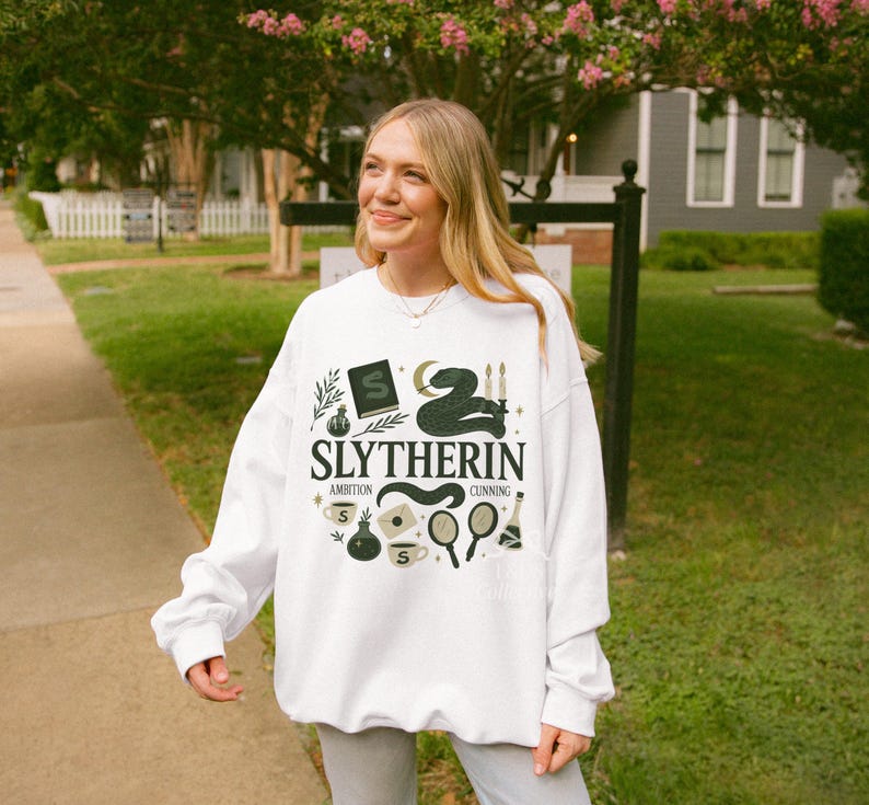 May include: A white long-sleeve sweatshirt featuring a dark green graphic print with the text SLYTHERIN, AMBITION, and CUNNING. The design includes illustrations of a snake, a book with an S, candles, potions, a mirror, and a coffee cup.