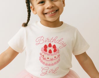 May include: A cream-colored t-shirt with a pink graphic of a birthday cake and the words "Birthday Girl." The cake has strawberries on top and is decorated with pink frosting and cherries. The girl is wearing a pink tutu.