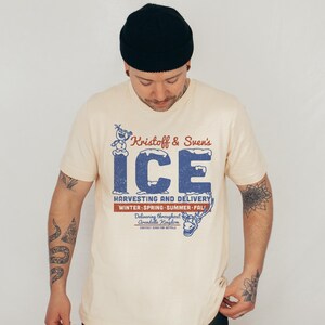 May include: Cream-colored t-shirt featuring a distressed blue graphic that reads "Kristoff & Sven's ICE" with an image of Olaf the snowman and Sven the reindeer. The shirt also includes text about harvesting and delivery.