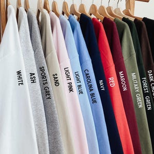 May include: A collection of thirteen crewneck sweatshirts hanging on wooden hangers, displayed in a color gradient from white to black. Each sweatshirt features printed text identifying its color: White, Ash, Sports Grey, Sand, Light Pink, Light Blue, Carolina Blue, Navy, Red, Maroon, Military Green, Forest Green, Dark Chocolate, and Black.