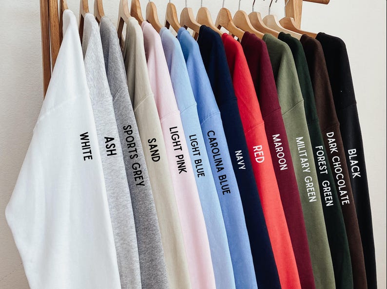May include: A collection of thirteen crewneck sweatshirts hanging on wooden hangers, displayed in a color gradient from white to black. Each sweatshirt features printed text identifying its color: White, Ash, Sports Grey, Sand, Light Pink, Light Blue, Carolina Blue, Navy, Red, Maroon, Military Green, Forest Green, Dark Chocolate, and Black.