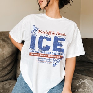 May include: White t-shirt featuring a distressed blue and red graphic. The design reads "Kristoff & Sven's ICE" with text about harvesting and delivery, and a reindeer illustration. The shirt is a casual, oversized fit.