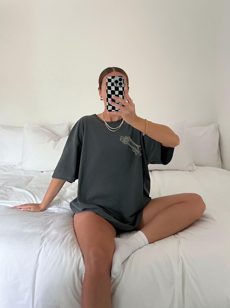 Pu&ograve; includere: Una persona indossa una maglietta oversize di colore grigio scuro con una stampa grafica di uno scheletro che tiene un fiore. La persona &egrave; seduta su un letto bianco con calzini bianchi.