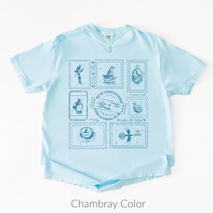 May include: Light blue t-shirt with a crew neck and a necklace. The shirt features a collection of blue postage stamp designs with travel-themed illustrations. The text "Travel" is visible on one of the stamps. The shirt is labeled "Chambray Color".