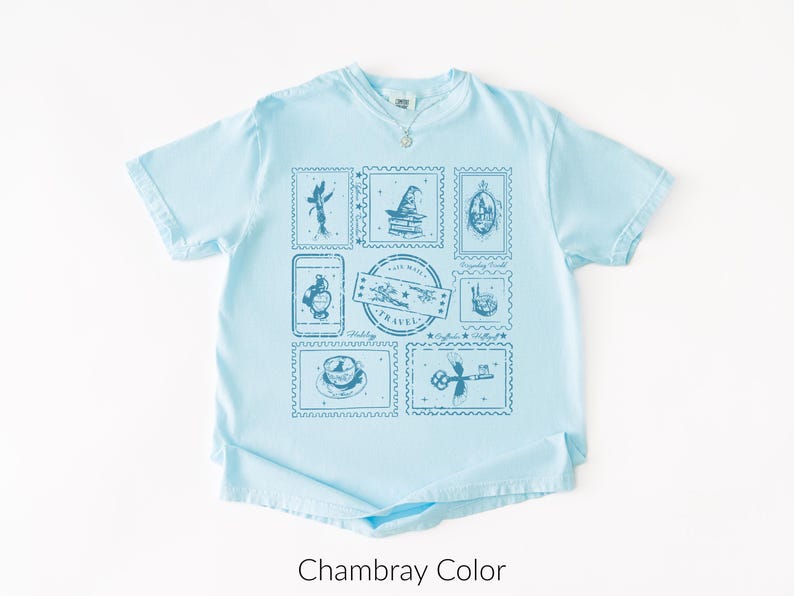 May include: Light blue t-shirt with a crew neck and a necklace. The shirt features a collection of blue postage stamp designs with travel-themed illustrations. The text "Travel" is visible on one of the stamps. The shirt is labeled "Chambray Color".