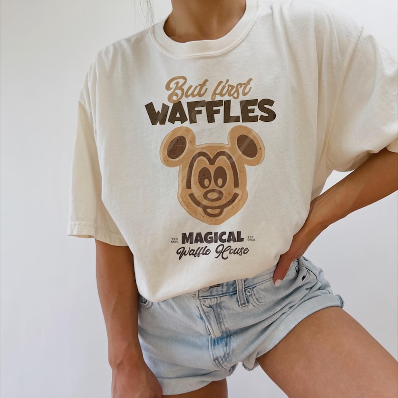 Puede incluir: Una camiseta blanca con un dise&ntilde;o de cabeza de Mickey Mouse marr&oacute;n y el texto "But first waffles" y "Magical Waffle House".