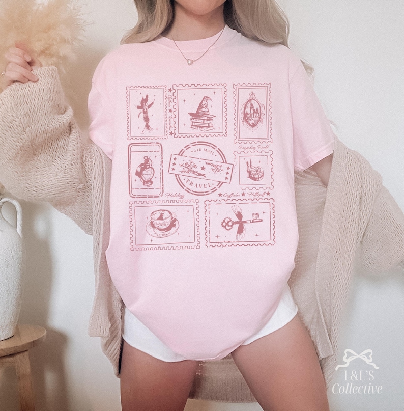 May include: A light pink t-shirt featuring a collection of postage stamp designs in a darker pink hue. The stamps depict various travel-themed illustrations, including a fairy, a wizard's hat, and a key. The words "Air Mail Travel" are also visible.