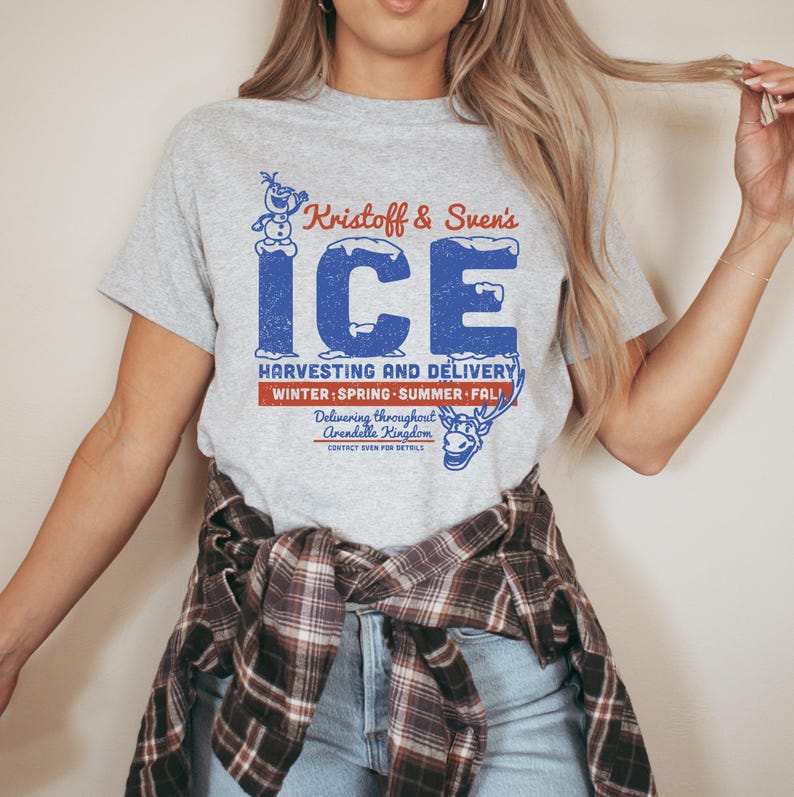 May include: A light gray t-shirt featuring a distressed blue graphic that reads "Kristoff & Sven's ICE." The design includes a snowman, a reindeer, and the text "Harvesting and Delivery" with seasonal references. The shirt is a crew neck.