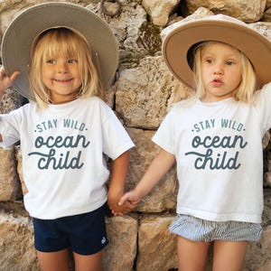 May include: Two children wearing white t-shirts with the text "Stay Wild. Ocean Child" in blue lettering. The children are wearing brown hats and holding hands.