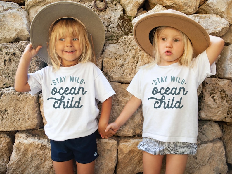 May include: Two children wearing white t-shirts with the text "Stay Wild. Ocean Child" in blue lettering. The children are wearing brown hats and holding hands.