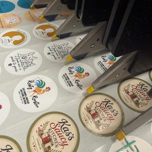 May include: A close-up of a machine applying labels to various round stickers. The stickers feature logos and text, including "Kass Saucy Kitchen" and "Rusty Rooster." The labels are in various colors and designs.