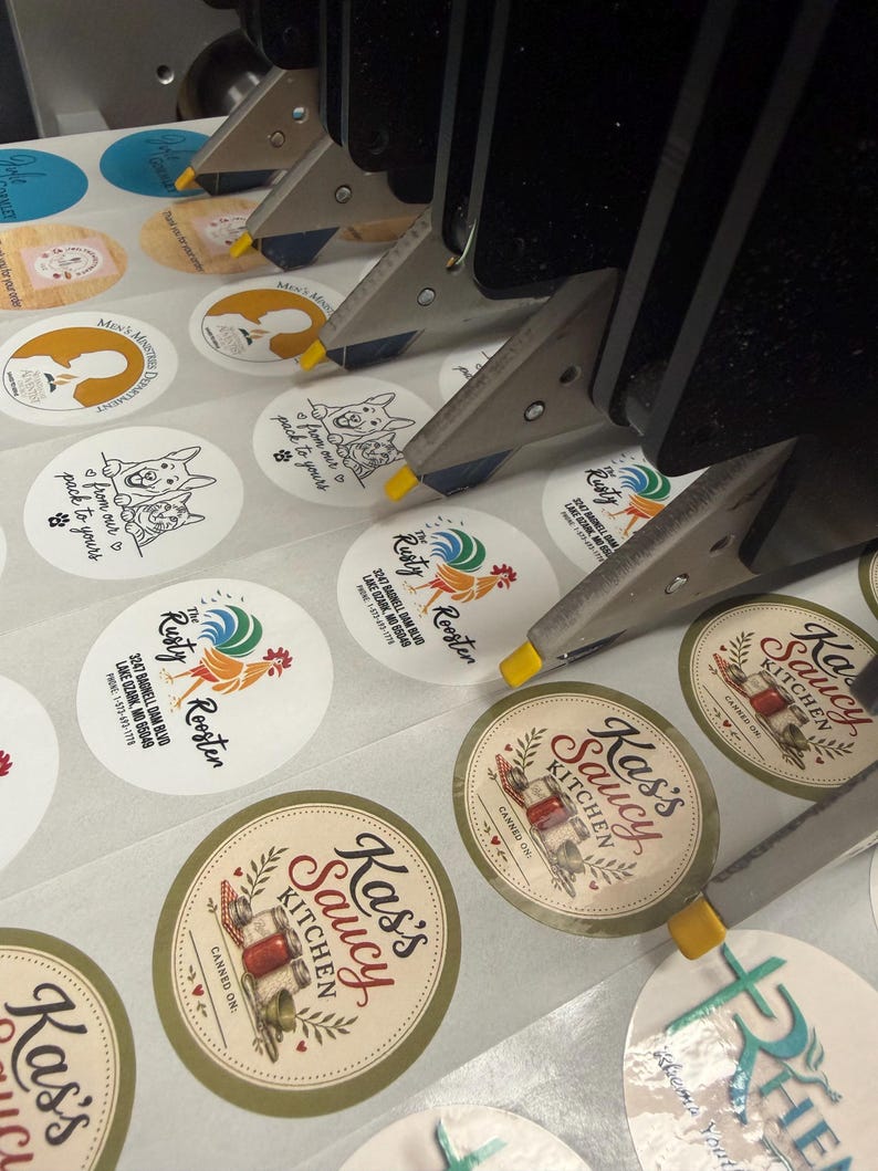 May include: A close-up of a machine applying labels to various round stickers. The stickers feature logos and text, including "Kass Saucy Kitchen" and "Rusty Rooster." The labels are in various colors and designs.