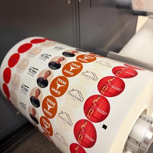 May include: A roll of printed circular stickers with various designs. Some stickers feature a person's portrait, others have the text "TEXAS DANCE" and a Longhorn logo, and some have a woman in a red dress. The stickers are in the process of being printed.
