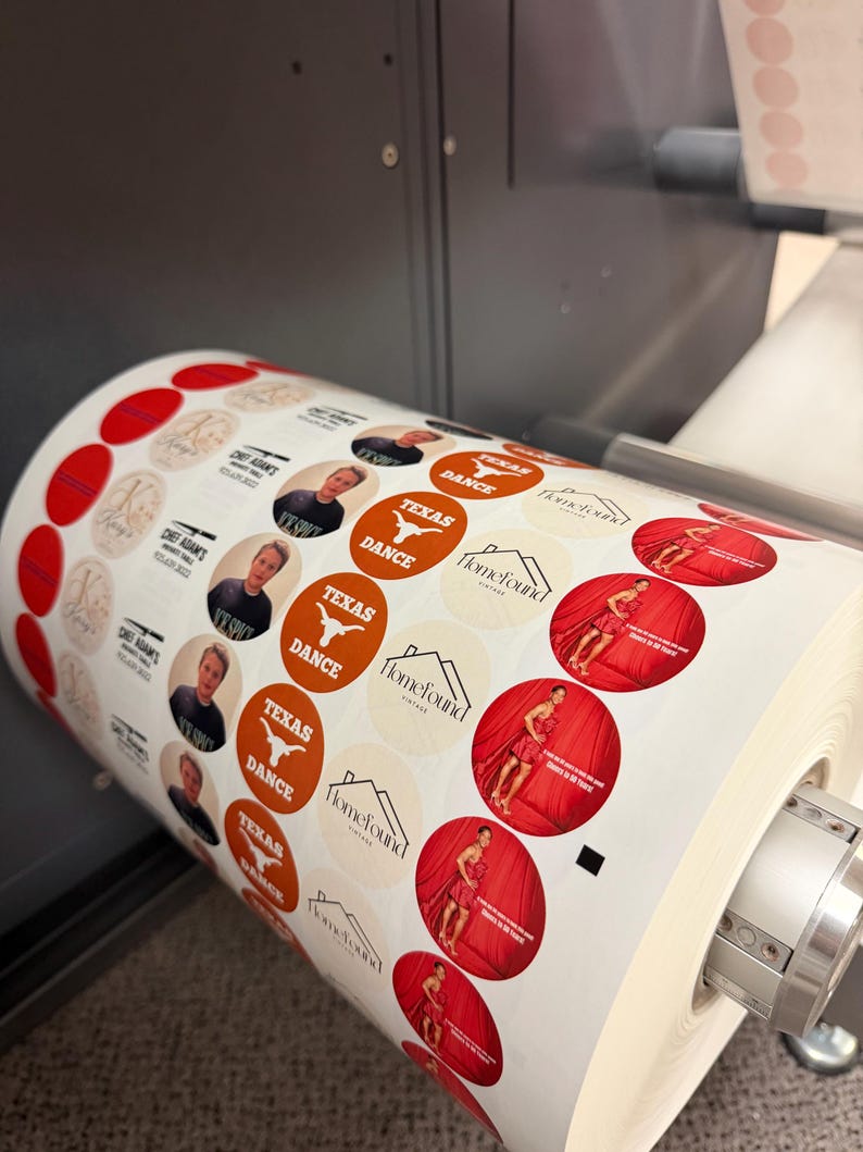May include: A roll of printed circular stickers with various designs. Some stickers feature a person's portrait, others have the text "TEXAS DANCE" and a Longhorn logo, and some have a woman in a red dress. The stickers are in the process of being printed.