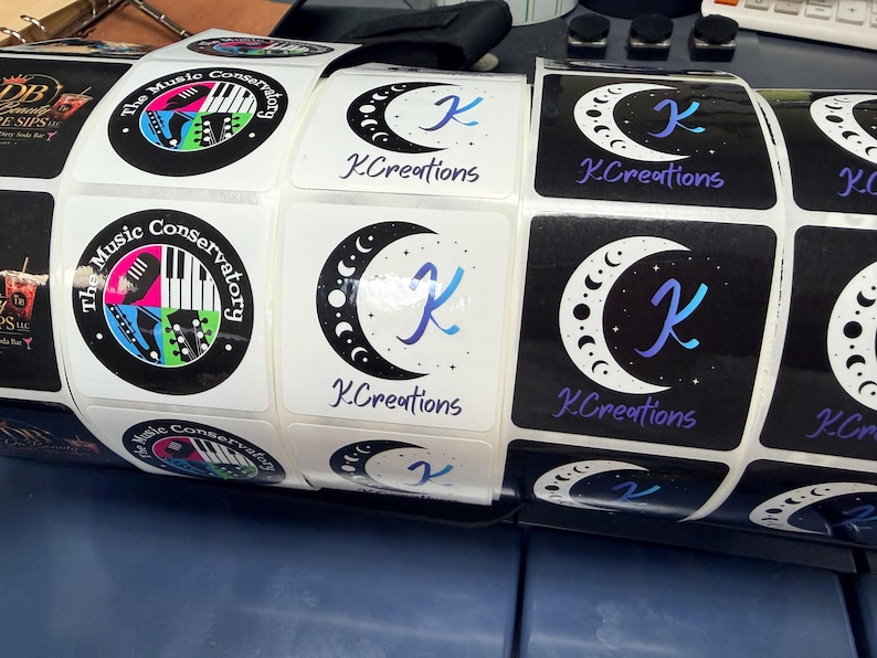 May include: Roll of square stickers featuring two designs. One has a black border with "The Music Conservatory" text and musical instrument graphics. The other has a crescent moon design with the letter "K" and "K.Creations" text.