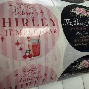May include: Roll of round stickers with pink and white striped backgrounds. The stickers feature the text "Aubrey's HIRLEY TEMPLE BAR" in burgundy, along with a cocktail illustration and cherries. Another sticker has the text "The Bizzy Butterfly".
