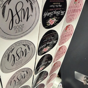 May include: A roll of printed stickers with various designs. Some stickers are oval-shaped with floral accents and the text "The Bizzy Butterfly." Others are round with "WSL 2005" and wheat illustrations. Pink and white striped stickers with the name "SHIRLEY TEMPLE BAR" are also visible.