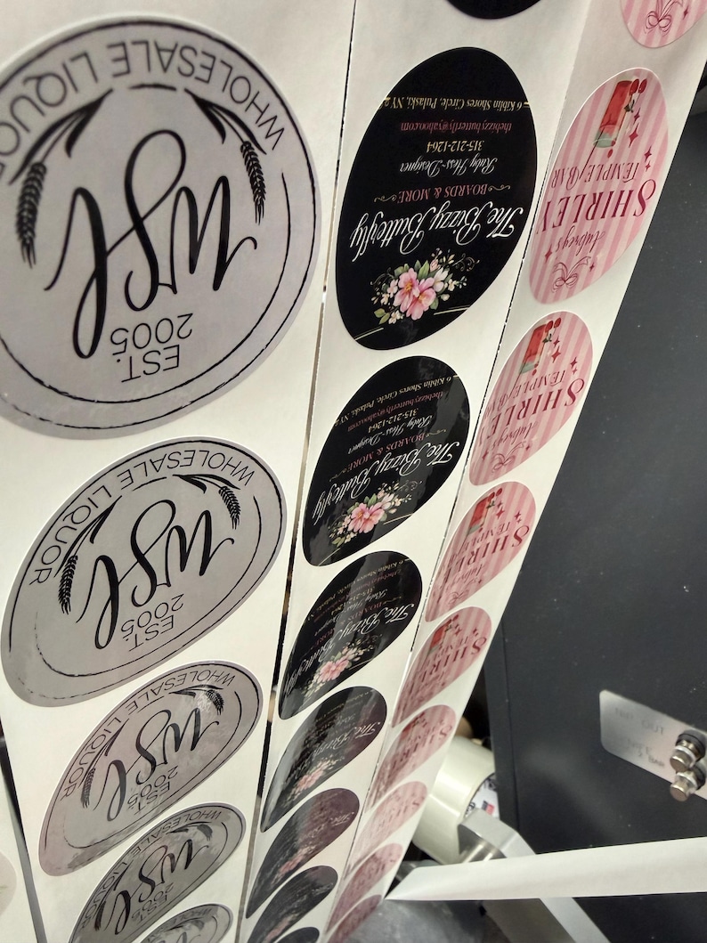 May include: A roll of printed stickers with various designs. Some stickers are oval-shaped with floral accents and the text "The Bizzy Butterfly." Others are round with "WSL 2005" and wheat illustrations. Pink and white striped stickers with the name "SHIRLEY TEMPLE BAR" are also visible.