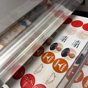 May include: A printing machine in operation, producing circular stickers. The stickers feature various designs, including the text "TEXAS DANCE", "Homefound Vintage", and images of people. Some stickers are red, orange, and beige.