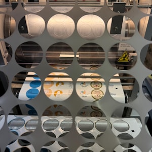 May include: A close-up view through a perforated metal sheet reveals a machine printing circular stickers. The stickers feature various designs and colors, including blue, yellow, and white. The machine has rollers and other mechanical components.
