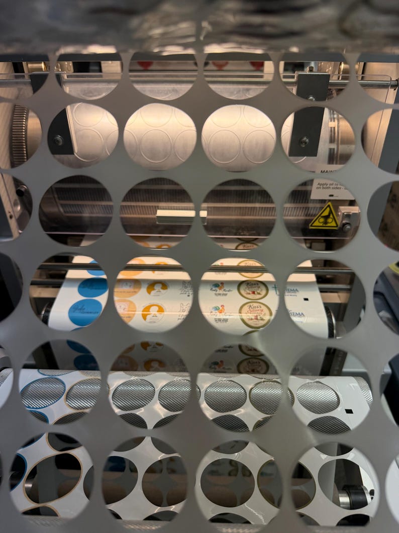 May include: A close-up view through a perforated metal sheet reveals a machine printing circular stickers. The stickers feature various designs and colors, including blue, yellow, and white. The machine has rollers and other mechanical components.